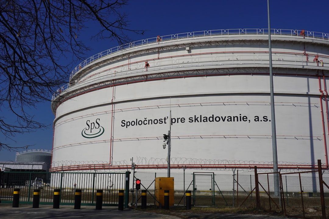 Wide landscape photo of large oil storage tanks at the Bučany petroleum storage facility in Slovakia, set behind a green field under a clear, pale blue sky. Large industrial storage tanks under a clear blue sky
